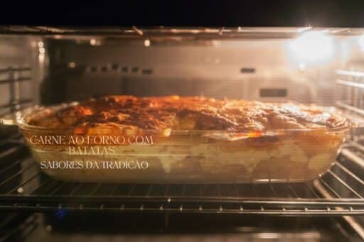 Uma assadeira com carne ao forno e batatas sendo cozida, vista através da porta de vidro de um forno iluminado.