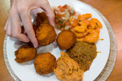 Uma mão pegando um acarajé de um prato com várias iguarias baianas, mostrando um típico prato da culinária da Bahia.