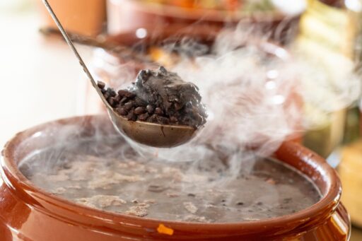 Uma concha servindo feijoada quente e fumegante de uma panela de barro, destacando a textura rica e o vapor aromático do prato.